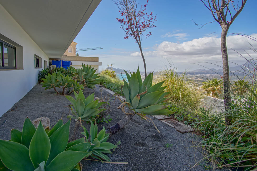 Der Seitengarten zeigt große Agaven auf dunklem Kies, angrenzend an eine weiße Fassade mit dunklen Fenstern. Er bietet Panoramablicke auf das Meer und ferne Berge.