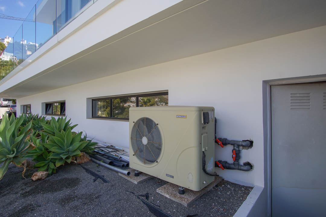 Der technische Außenbereich der Villa zeigt eine AstralPool Inverter-Wärmepumpe, horizontale Fenster und eine Versorgungstür. Agaven schmücken die moderne weiße Fassade.