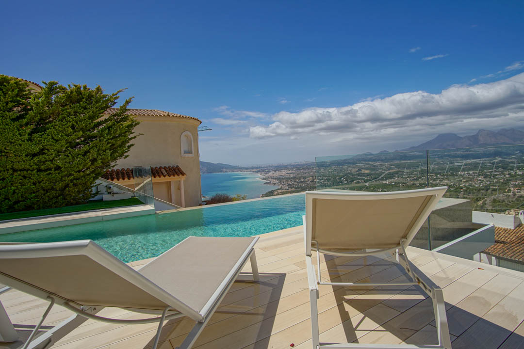 Die Außenterrasse verfügt über einen Infinity-Pool mit Blick auf die Küste und ferne Berge. Zwei Sonnenliegen auf hellem Deck, eingerahmt von einer Glasbrüstung und einer traditionellen Villenfassade.