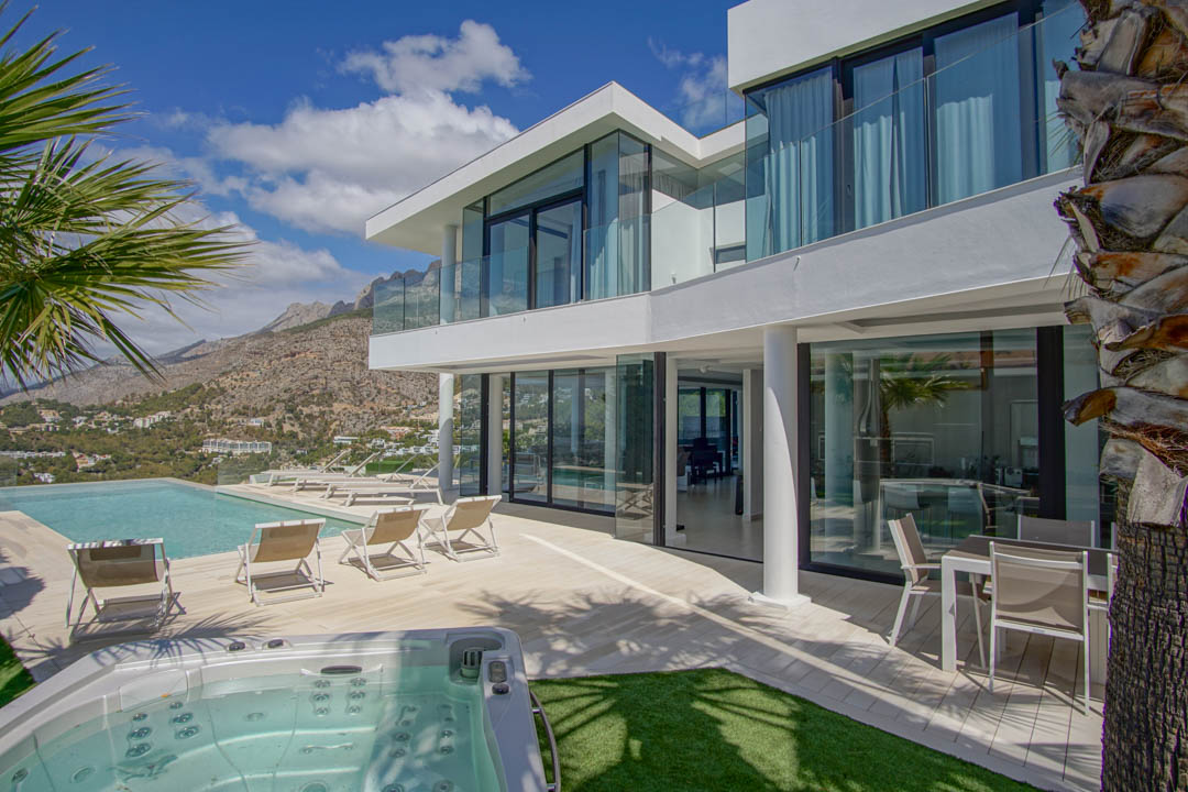 Die moderne weiße Villa außen zeigt einen Infinity-Pool, separaten Whirlpool und eine großzügige Terrasse mit Liegen, umrahmt von Bergpanoramen unter klarem Himmel.