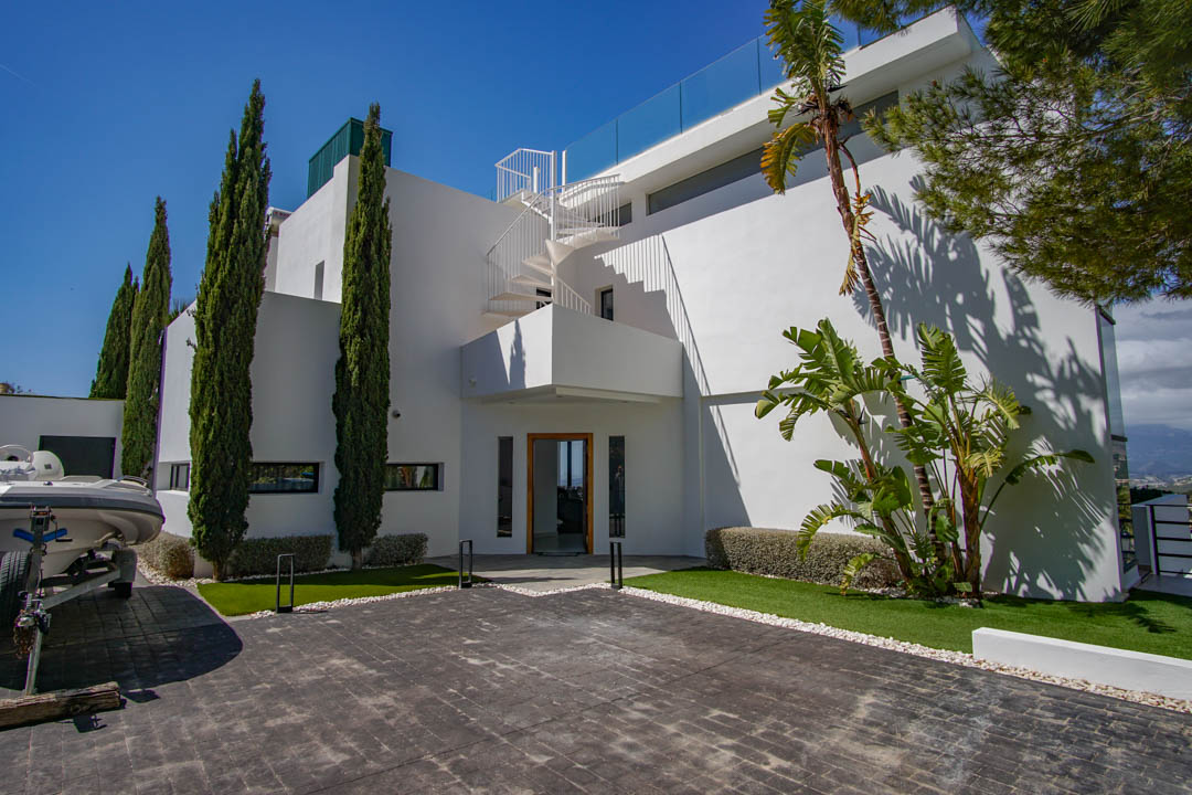 Die moderne weiße Villa zeigt eine gepflasterte Auffahrt, hohe Zypressen und eine markante Wendeltreppe, die zur oberen Terrasse mit Glasgeländer führt.