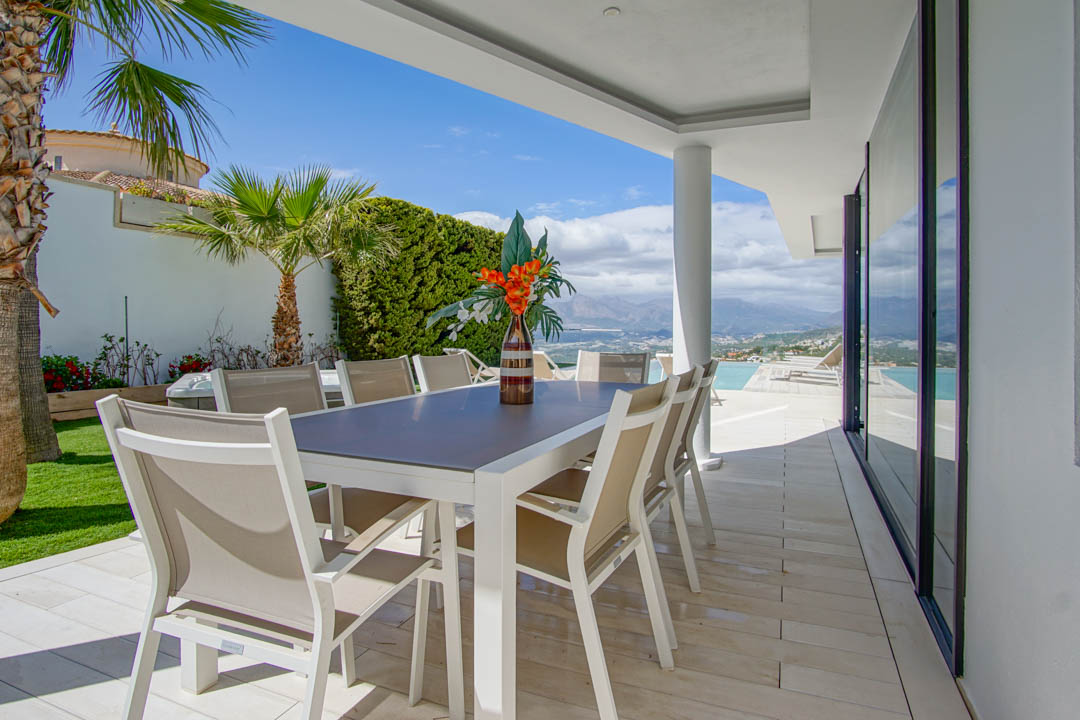 Diese überdachte Außenterrasse verfügt über einen modernen weißen Tisch mit acht beigen Stühlen auf hellen Fliesen. Sie bietet weite Ausblicke auf die Berge und das Meer, angrenzend an den Infinity-Pool.