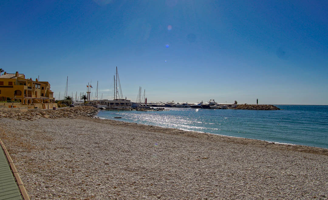 Ein breiter Kieselstrand erstreckt sich zu einem Yachthafen voller Yachten, gesäumt von traditionellen Küstengebäuden unter klarem, blauem Himmel.