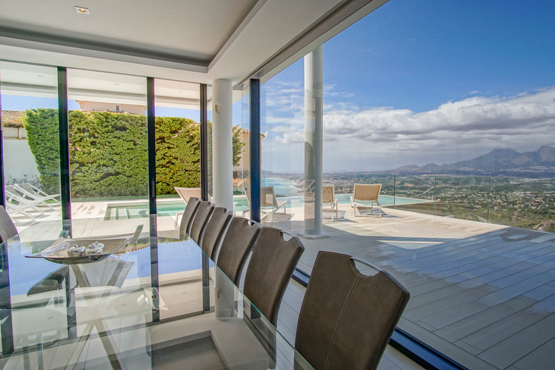Eleganter Essbereich im Innenbereich mit Glastisch und Stühlen, der nahtlos in eine weitläufige Terrasse mit Infinity-Pool und Panoramablick auf Meer und Berge übergeht.