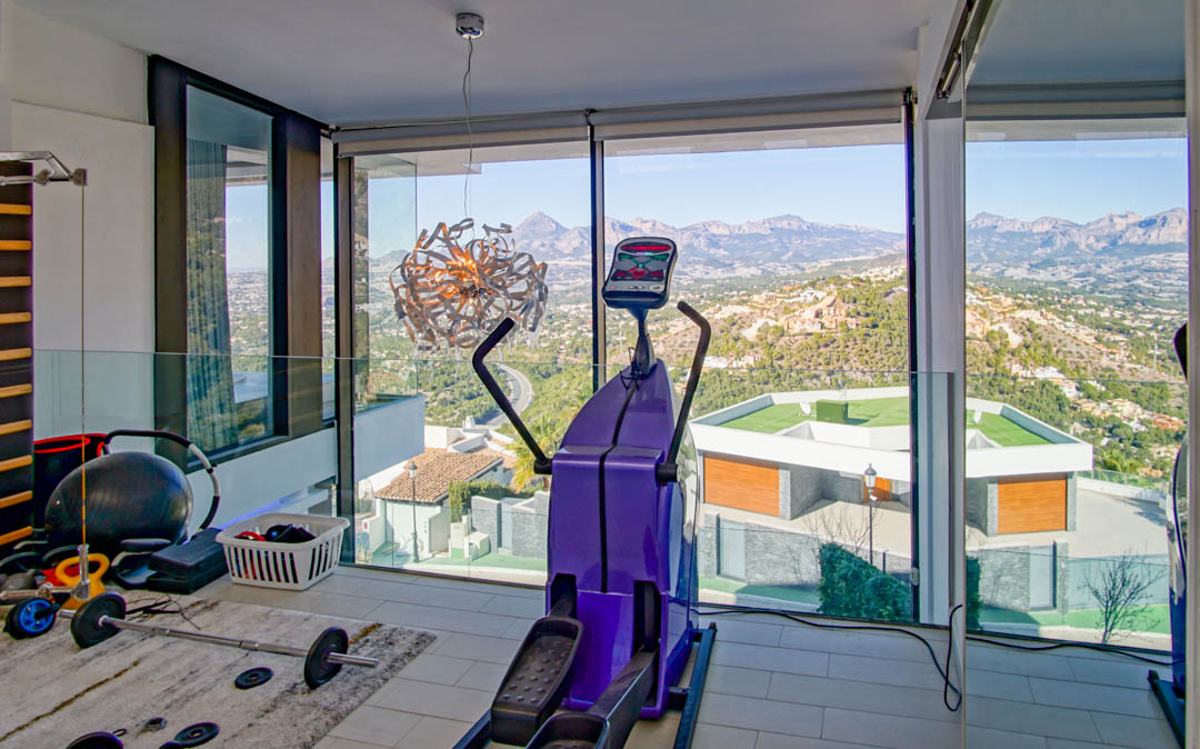 Fitnessraum mit raumhohen Fenstern, die einen Panoramablick auf Berge und Tal bieten. Ausgestattet mit Ellipsentrainer und Gewichten, ergänzt durch eine Glasbalustrade.