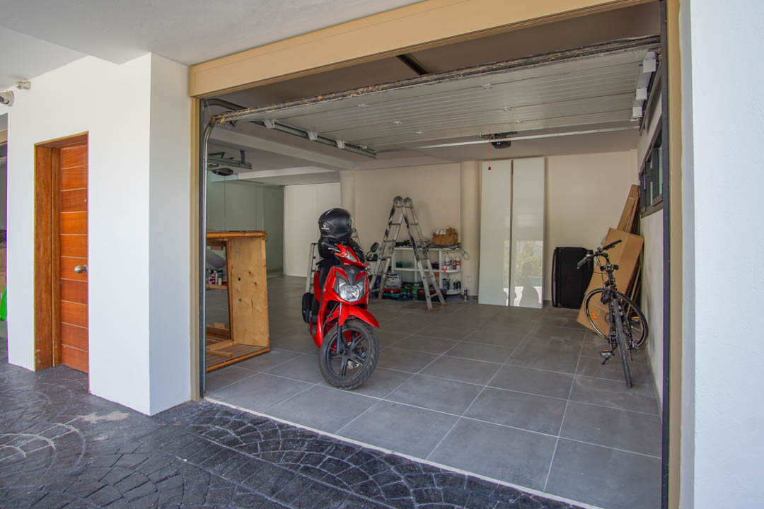 Geräumige Garage mit grauem Fliesenboden, einem teilweise geöffneten Sektionaltor und einer markanten Holzeingangstür links. Ein roter Roller und ein Fahrrad sind sichtbar.