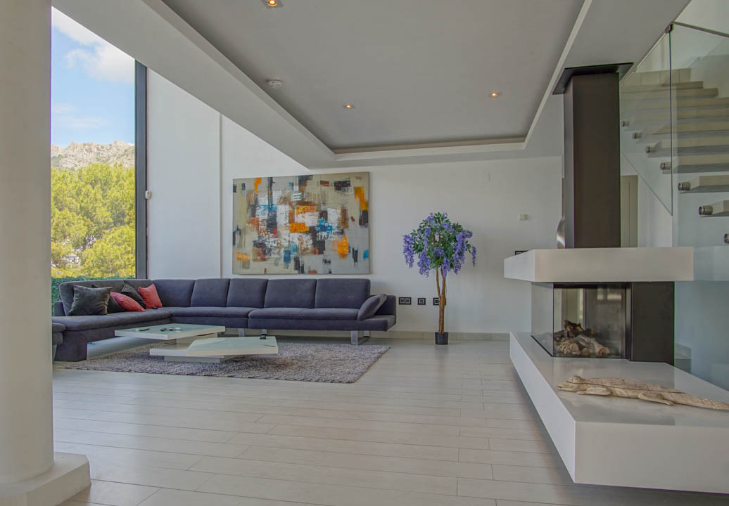 Geräumiges Wohnzimmer mit grauem Ecksofa, modernen Couchtischen und Gaskamin. Große Fenster bieten Berg- und Baumblick, ergänzt durch eine Glastreppe.
