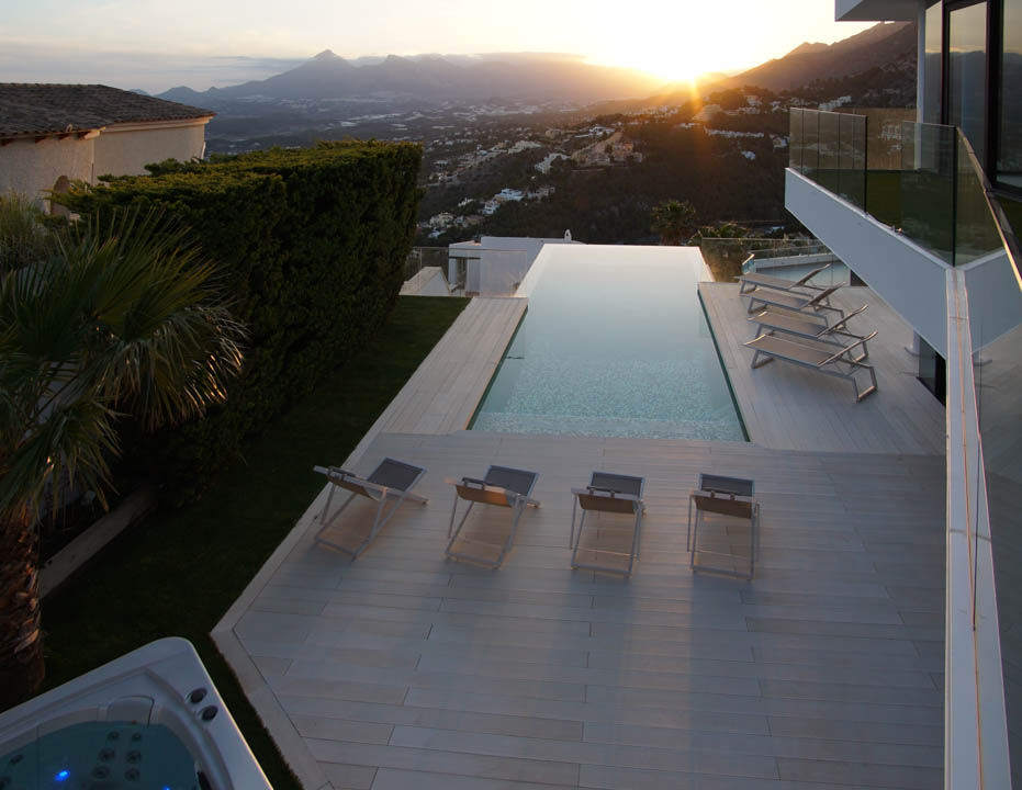 Großer Infinity-Pool mit heller Holzterrasse, mehreren Sonnenliegen und separatem Whirlpool, umrahmt von einer Gebirgslandschaft bei Sonnenuntergang.