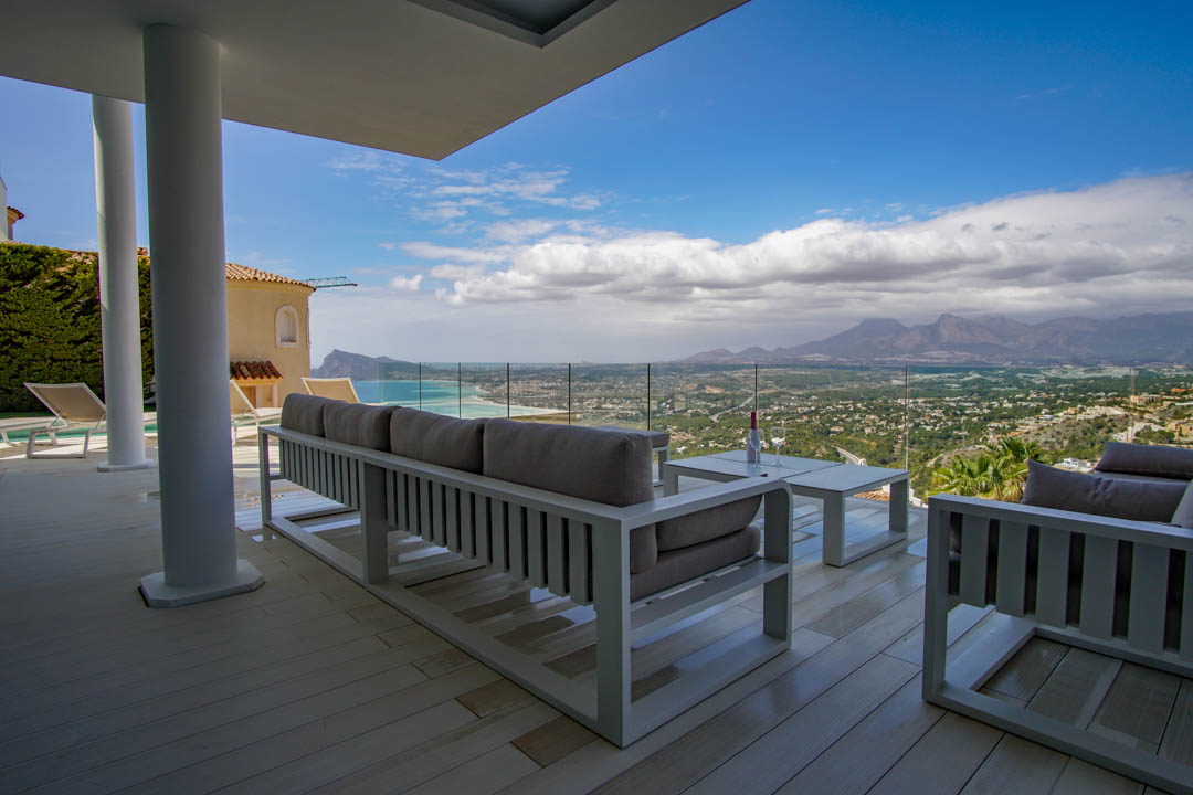 Großzügige überdachte Terrasse mit hellem Bodenbelag und weißen Loungemöbeln, die durch eine Glasbrüstung einen Panoramablick auf Meer, Küste und Berge bietet.