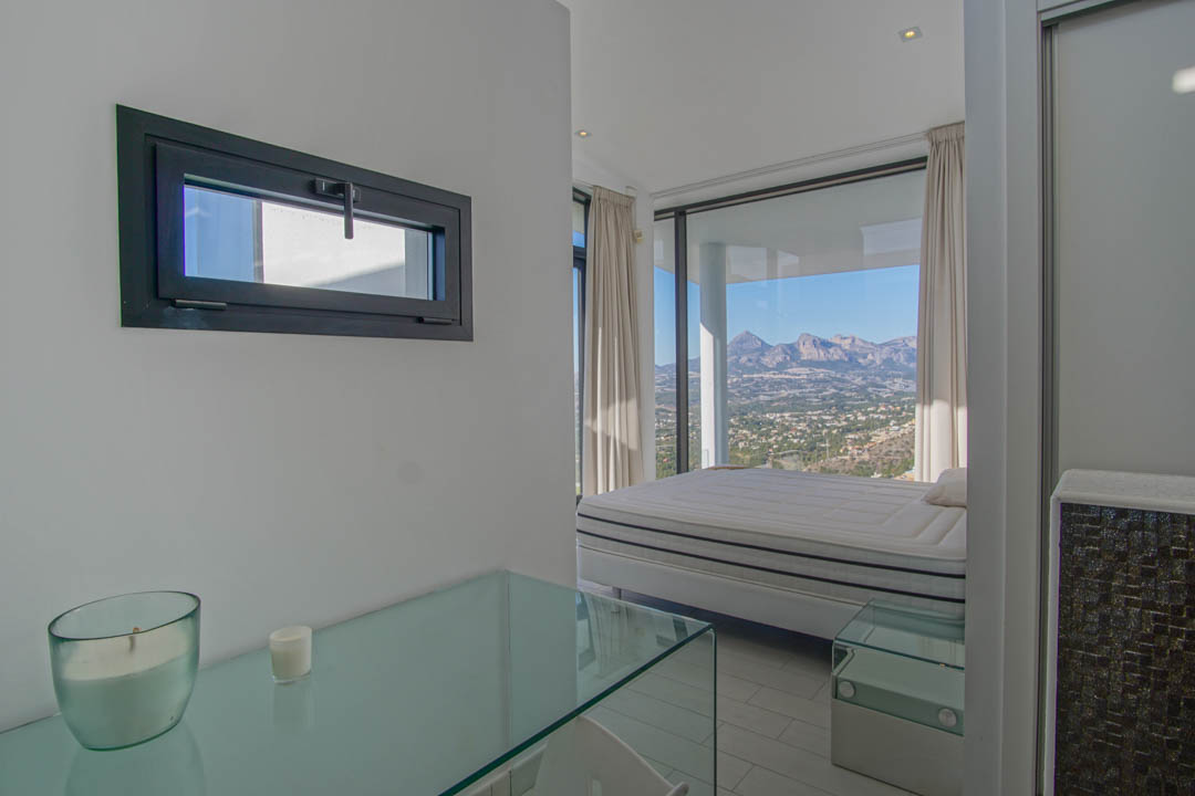Helles Schlafzimmer mit Bett vor raumhohen Fenstern, die einen Panoramablick auf Berge und Tal bieten. Eine Glaskonsole und ein horizontales Fenster mit dunklem Rahmen sind ebenfalls sichtbar.