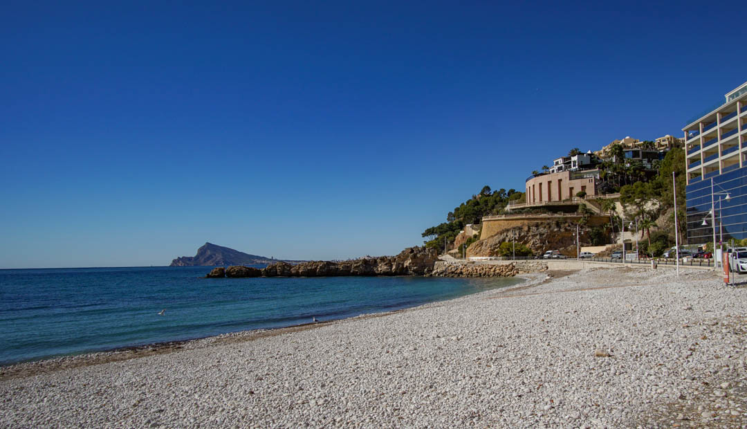 Kieselstrand mit klarem türkisfarbenem Wasser und felsiger Küste. Moderne Gebäude mit reflektierenden Fassaden und ein Rundbau säumen den Hang unter einem strahlend blauen Himmel.