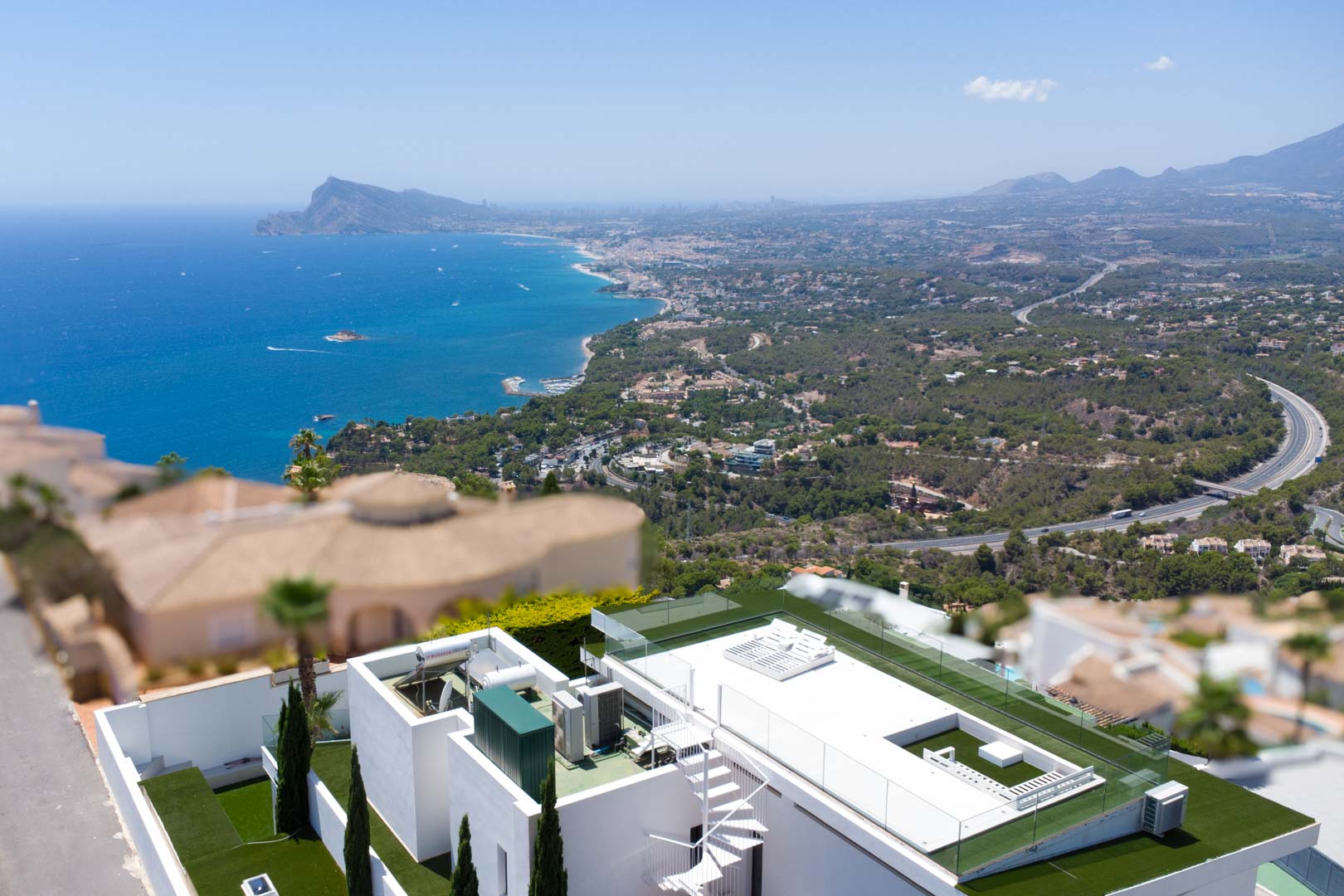 Luftaufnahme der Dachterrasse der Villa mit weißer Wendeltreppe, Kunstrasen, Glasgeländer und weitem Blick auf das Mittelmeer und die Küstenlandschaft.