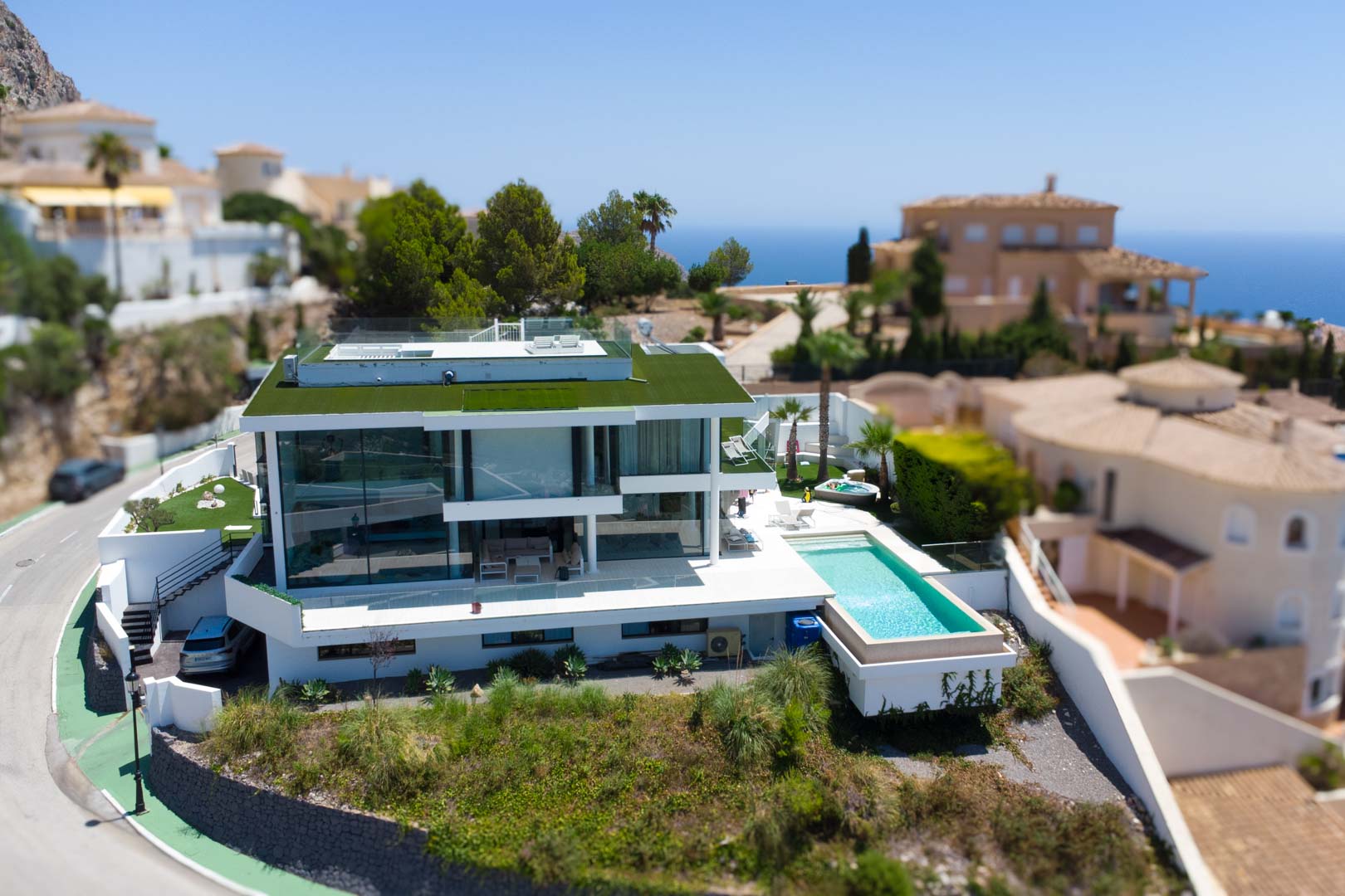 Luftaufnahme der modernen weißen Designervilla mit Gründach, Infinity-Pool und separatem Whirlpool, eingebettet in eine Umgebung mit Meerblick.