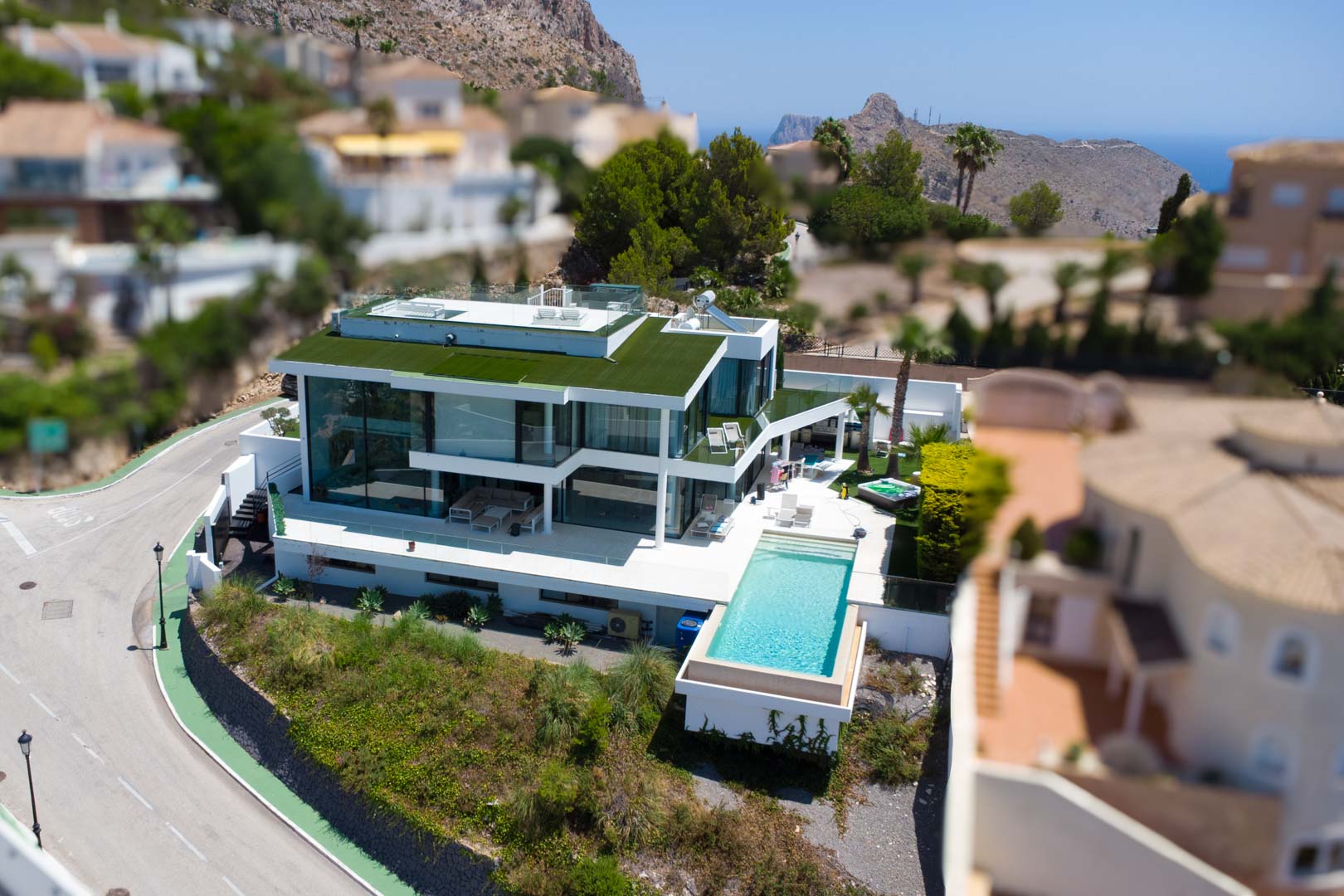 Luftaufnahme einer modernen weißen Villa mit Infinity-Pool, großen Glasfronten, begrüntem Dach und separatem Whirlpool, mit Berg- und Meerblick.