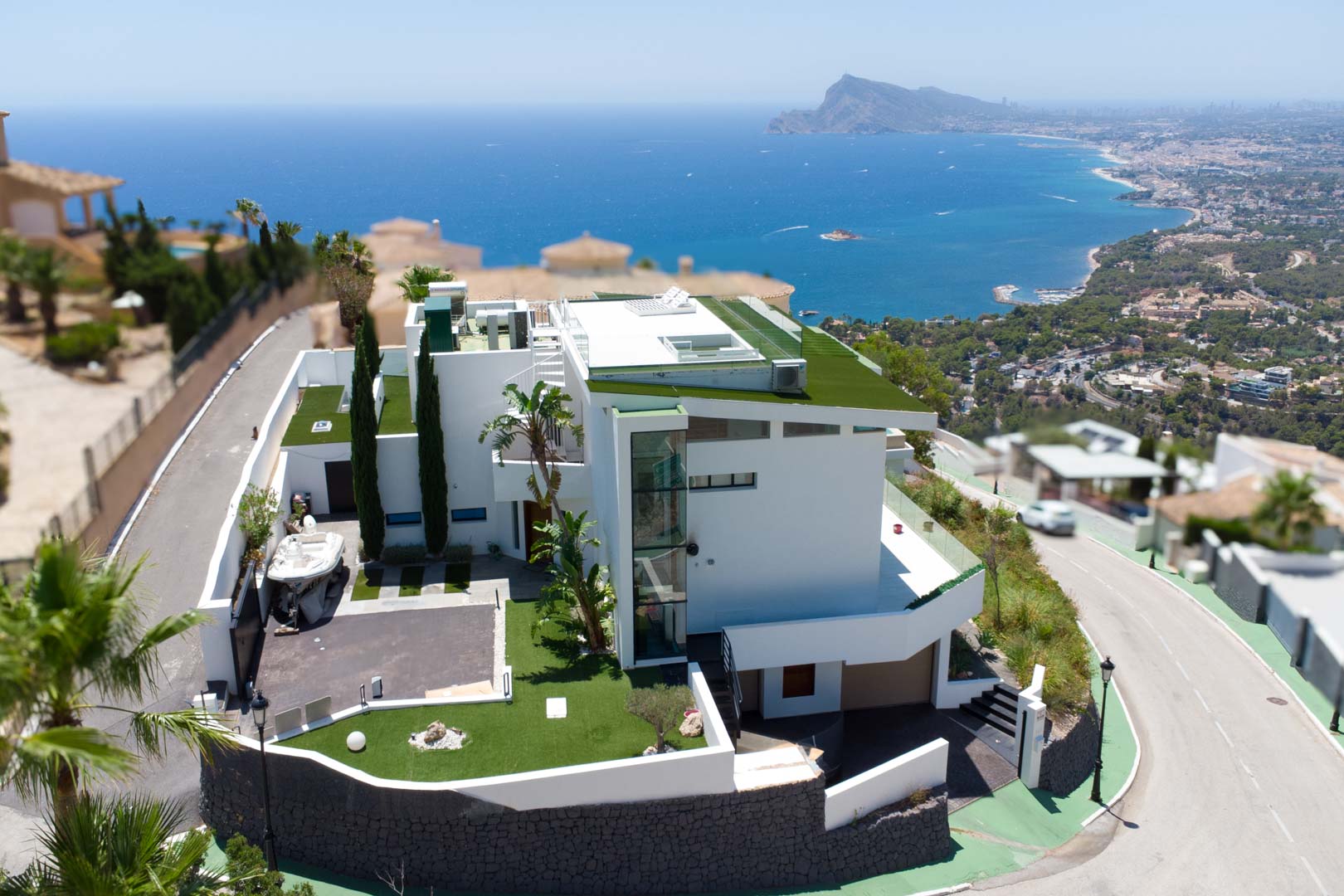 Luftaufnahme einer modernen weißen Villa mit mehrstufiger Architektur, begrünten Flachdächern und externem Glasaufzug. Das Anwesen umfasst eine private Auffahrt und bietet Panorama-Meer- und Küstenblicke.