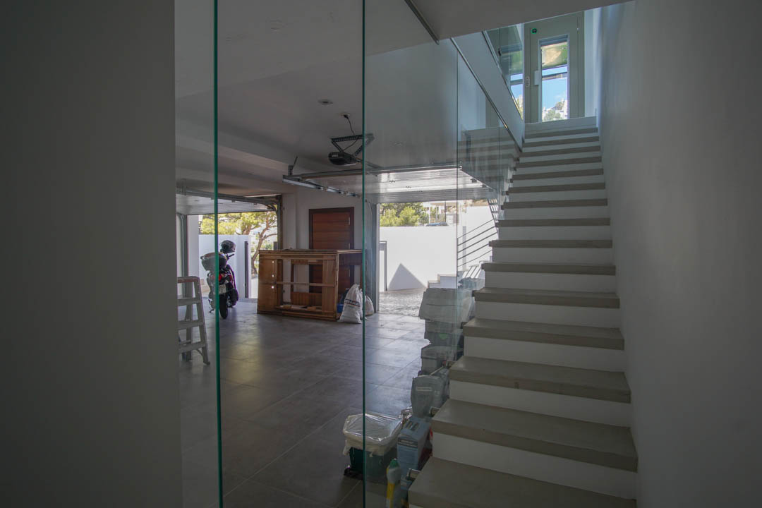 Moderne Innentreppe mit Glasgeländer und hellen Stufen, angrenzend an eine geflieste Garage mit offenem Tor und natürlichem Lichteinfall.
