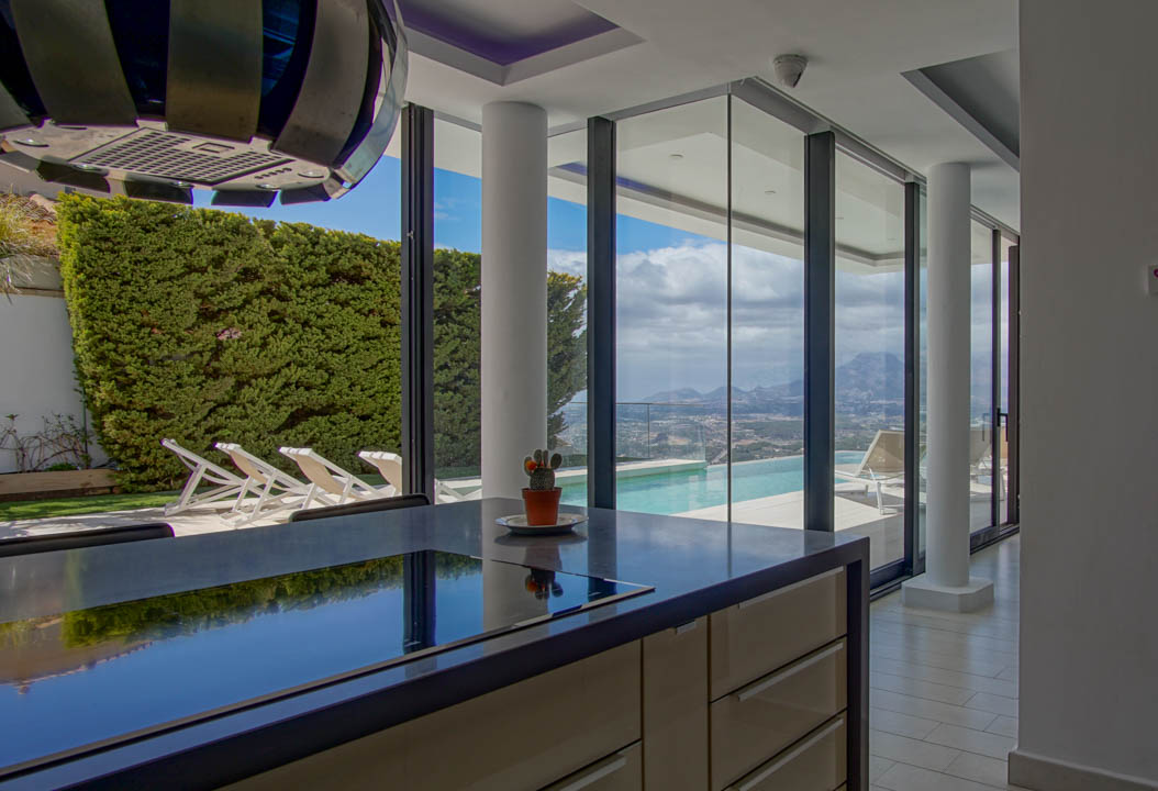 Moderne Kücheninsel mit dunkler Arbeitsplatte und integriertem Kochfeld. Die bodentiefe Fensterfront bietet einen weiten Blick auf den Infinity-Pool, das Tal und die Berge.