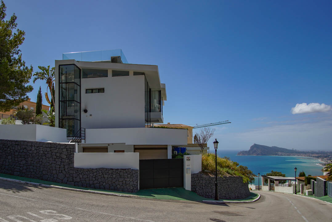 Moderne weiße Villa mit sichtbarem Glasaufzug und Dachterrassen-Brüstung. Steinmauern und ein automatisches Tor ergänzen den Panoramablick auf das Meer und die Küste.