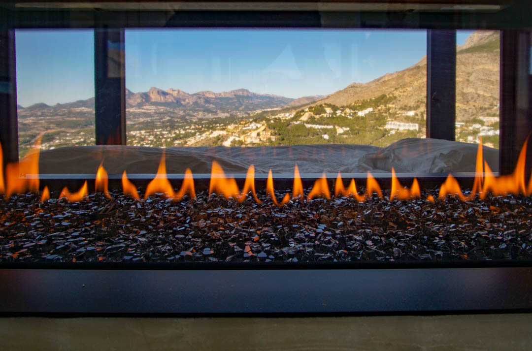 Moderner Gaskamin mit Flammen über dunklem Glasgranulat, der einen Panoramablick auf Berge und Tal freigibt.