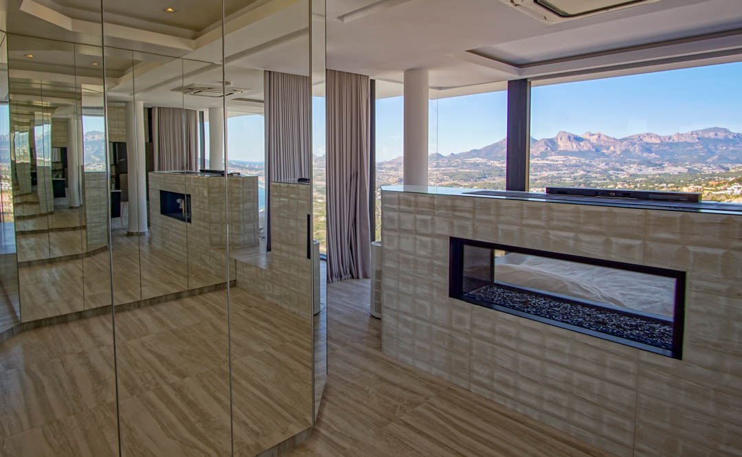Moderner Innenraum mit transparentem Gaskamin und Kiesbett, in eine strukturierte Wand integriert. Spiegelpaneele reflektieren den Raum und die weiten Berg- und Meerblicke durch raumhohe Fenster.