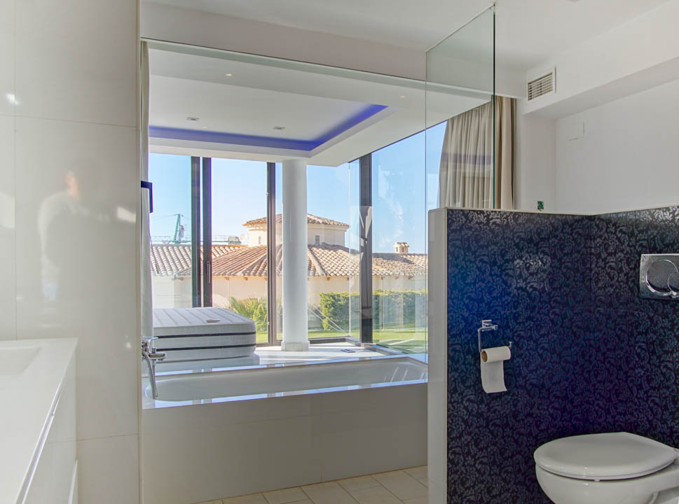 Modernes Badezimmer mit integrierter Badewanne und raumhohen Fenstern, die Blick auf ein Ziegeldach und den Garten bieten. Blaue Akzentbeleuchtung und eine dunkel gemusterte Trennwand sind vorhanden.