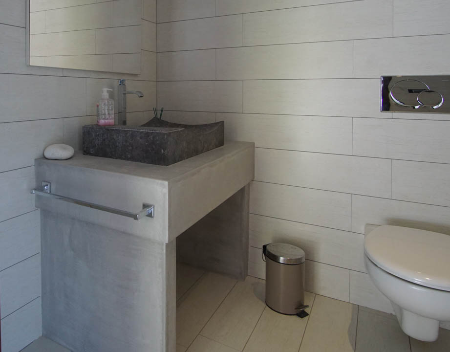 Modernes Gäste-WC mit Steinwaschbecken auf Betonoptik-Waschtisch und hellen Fliesen. Wandhängendes WC mit Chrom-Spülplatte.