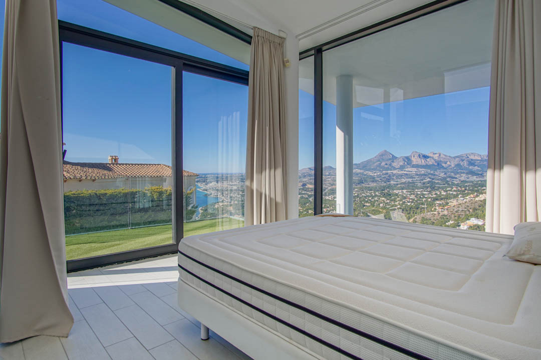 Modernes Schlafzimmer mit großem Bett und weiten Fenstern. Es bietet Panoramablicke auf das ferne Meer, eine grüne Rasenfläche und schroffe Berge unter klarem Himmel.