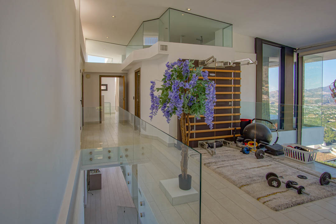 Obergeschoss-Fitnessbereich mit modernen Glasgeländern und hellem Boden. Ausgestattet mit Sprossenwand und Gewichten, bietet er Panoramablicke durch raumhohe Fenster.