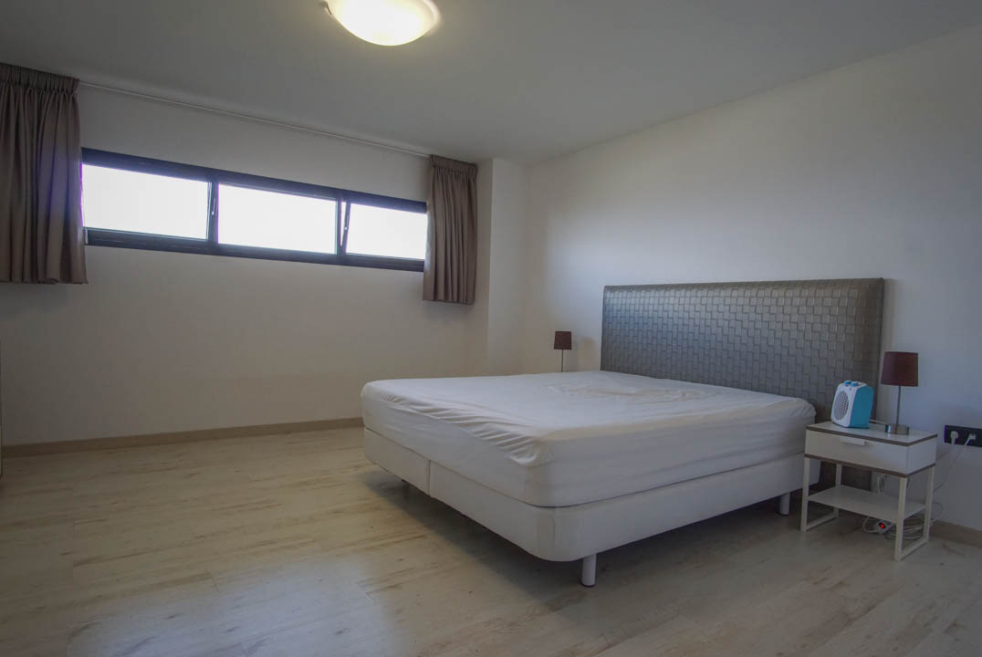 Schlafzimmer mit hellem Holzfußboden und weißen Wänden. Es verfügt über ein Bett mit strukturiertem Kopfteil, Nachttisch und ein horizontales Fenster mit dunklen Rahmen.