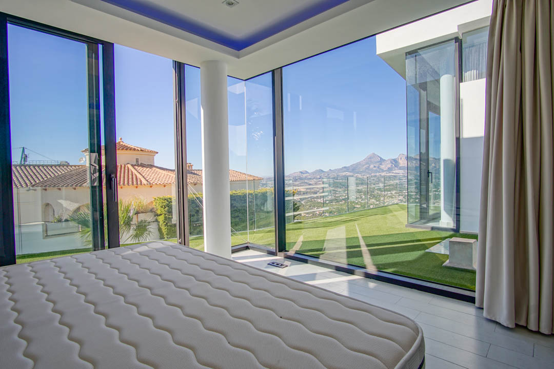 Schlafzimmer mit raumhohen Eckglaswänden, das einen Panoramablick auf ferne Berge, ein Tal und benachbarte Dächer bietet. Ein weißer Stützpfeiler und direkter Zugang zu Kunstrasen sind vorhanden.