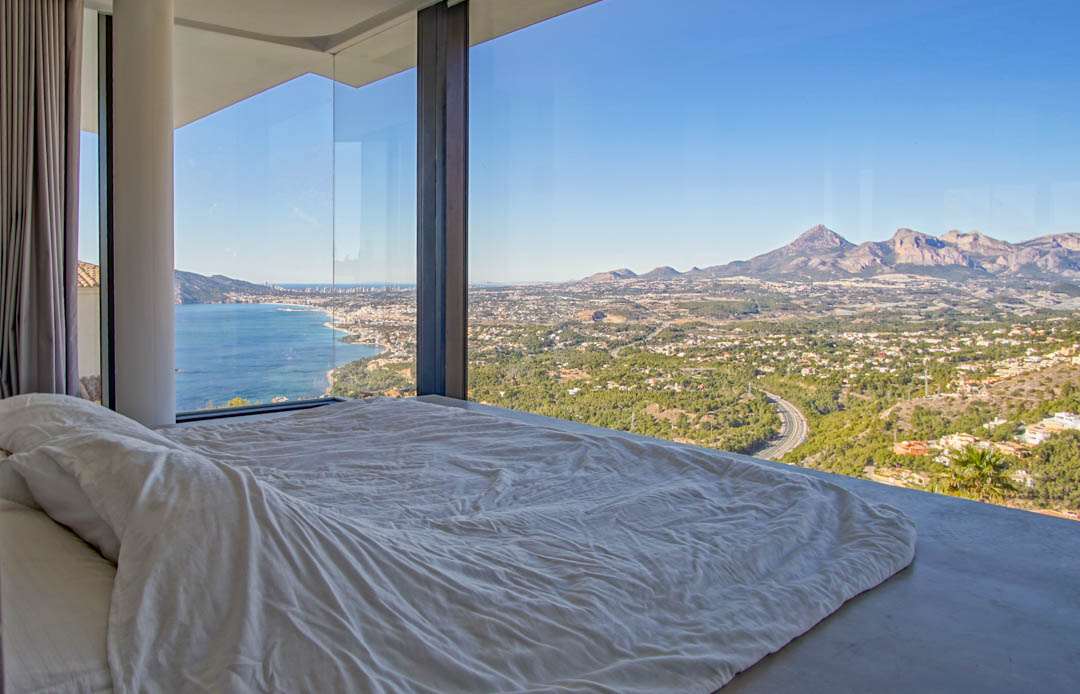 Schlafzimmer mit raumhohen Glasfenstern und Panoramablick auf das Mittelmeer, eine Küstenstadt und ferne Berge. Ein niedriges Bett mit weißer Bettwäsche orientiert sich zur Aussicht.