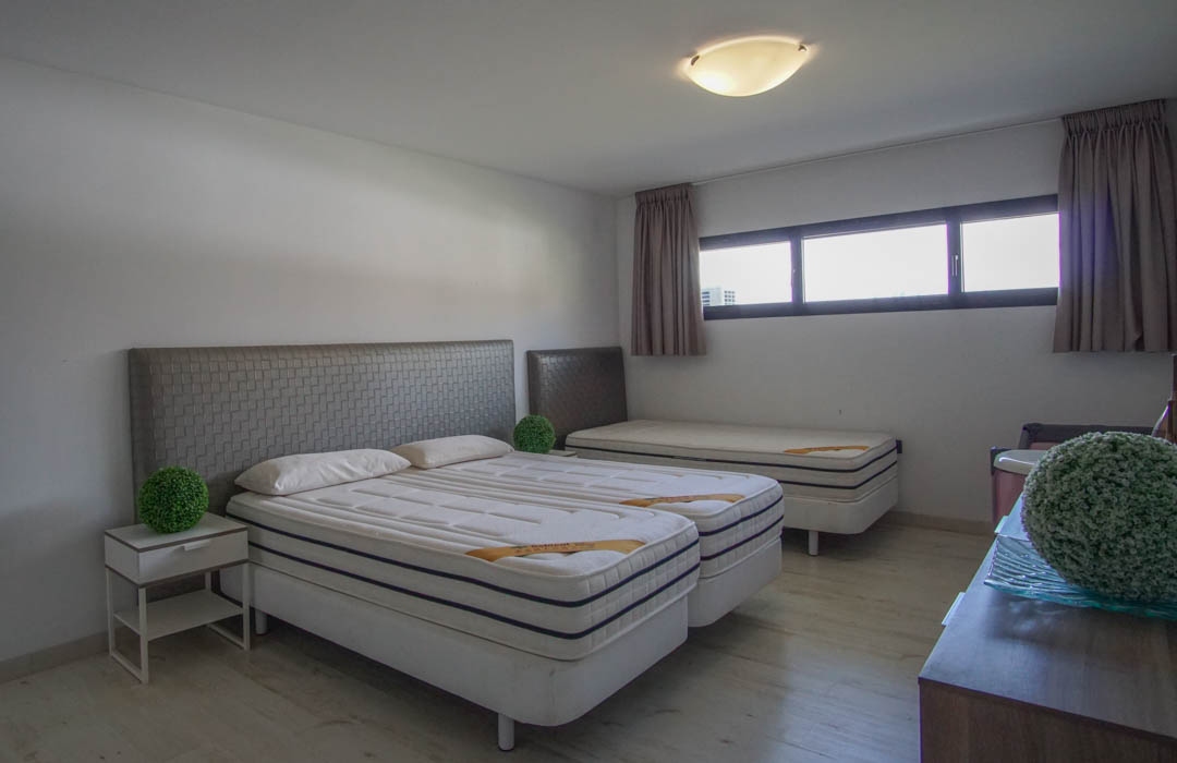 Schlafzimmer mit zwei kombinierten Einzelbetten, grauen strukturierten Kopfteilen, einem dritten Einzelbett im Hintergrund, hellem Holzboden und horizontalen Fenstern.