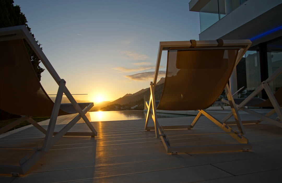 Zwei moderne Liegestühle auf heller Terrasse blicken auf einen Infinity-Pool, der den goldenen Sonnenuntergang über den Bergen spiegelt.