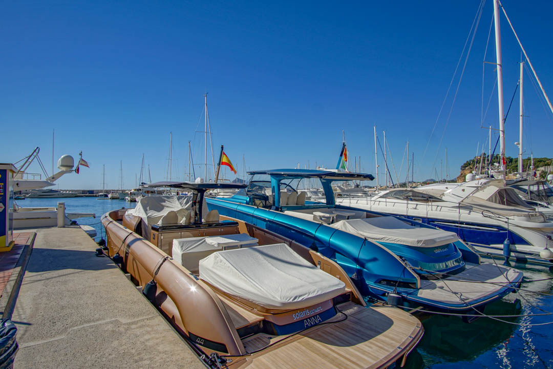 Zwei moderne Motorboote, eines bronze mit Teakdeck und ein weiteres metallicblau, liegen in einem Yachthafen unter klarem Himmel. Spanische Flaggen sind unter den Yachten im Hintergrund zu sehen.