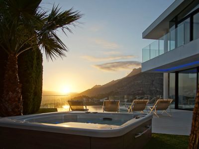 Außen-Jacuzzi mit Holzoptik auf Rasenfläche, vor einer modernen Villa mit Glasbalkonen. Panoramablick auf Berge und Meer bei Sonnenuntergang.