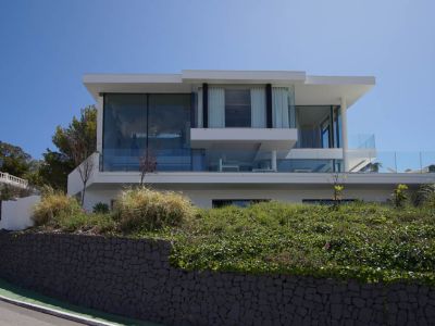 Außenansicht einer modernen dreistöckigen Villa mit weitläufigen Glasfassaden und Glasgeländern, erhöht über einer Steinmauer und Vegetation an einer geschwungenen Straße.