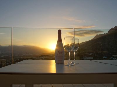 Außentisch auf einer Terrasse mit Glasgeländer, mit Rosé-Cava-Flasche und zwei Gläsern. Sonnenuntergang über Bergen und Tal, mit warmen Reflexionen des Abendlichts.