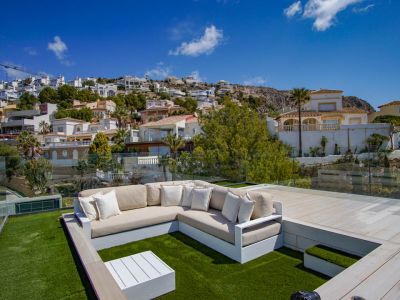 Dachterrassen-Lounge mit L-förmigem Sofa auf Kunstrasen, weißem Couchtisch und Glasgeländer. Bietet klare Aussicht auf den terrassierten Hang mit Wohnhäusern.