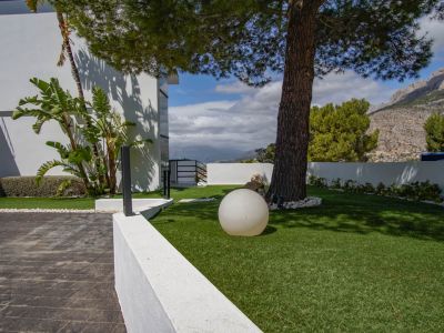 Der Außenbereich der Villa zeigt eine gepflasterte Auffahrt, Kunstrasen mit einer alten Kiefer, eine moderne Kugelleuchte und Bergblick.