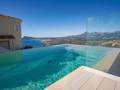 Der Infinity-Pool mit klarem Wasser, heller Decking und Glasbarriere bietet einen Panoramablick auf Meer, Küste und ferne Berge unter blauem Himmel.