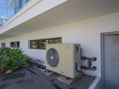 Der technische Außenbereich der Villa zeigt eine AstralPool Inverter-Wärmepumpe, horizontale Fenster und eine Versorgungstür. Agaven schmücken die moderne weiße Fassade.