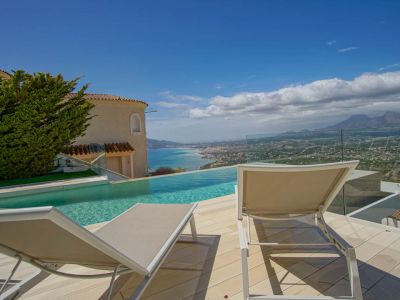 Die Außenterrasse verfügt über einen Infinity-Pool mit Blick auf die Küste und ferne Berge. Zwei Sonnenliegen auf hellem Deck, eingerahmt von einer Glasbrüstung und einer traditionellen Villenfassade.