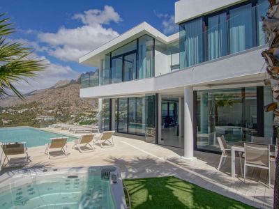 Die moderne weiße Villa außen zeigt einen Infinity-Pool, separaten Whirlpool und eine großzügige Terrasse mit Liegen, umrahmt von Bergpanoramen unter klarem Himmel.