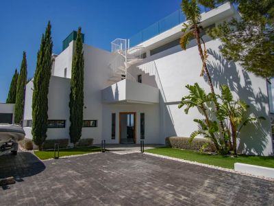 Die moderne weiße Villa zeigt eine gepflasterte Auffahrt, hohe Zypressen und eine markante Wendeltreppe, die zur oberen Terrasse mit Glasgeländer führt.
