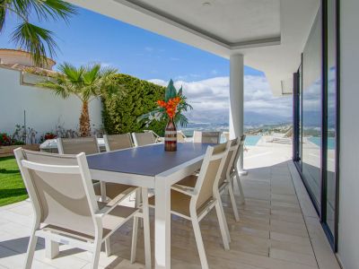 Diese überdachte Außenterrasse verfügt über einen modernen weißen Tisch mit acht beigen Stühlen auf hellen Fliesen. Sie bietet weite Ausblicke auf die Berge und das Meer, angrenzend an den Infinity-Pool.
