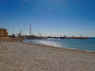 Ein breiter Kieselstrand erstreckt sich zu einem Yachthafen voller Yachten, gesäumt von traditionellen Küstengebäuden unter klarem, blauem Himmel.