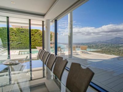 Eleganter Essbereich im Innenbereich mit Glastisch und Stühlen, der nahtlos in eine weitläufige Terrasse mit Infinity-Pool und Panoramablick auf Meer und Berge übergeht.