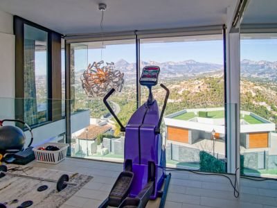 Fitnessraum mit raumhohen Fenstern, die einen Panoramablick auf Berge und Tal bieten. Ausgestattet mit Ellipsentrainer und Gewichten, ergänzt durch eine Glasbalustrade.