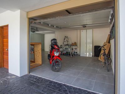 Geräumige Garage mit grauem Fliesenboden, einem teilweise geöffneten Sektionaltor und einer markanten Holzeingangstür links. Ein roter Roller und ein Fahrrad sind sichtbar.