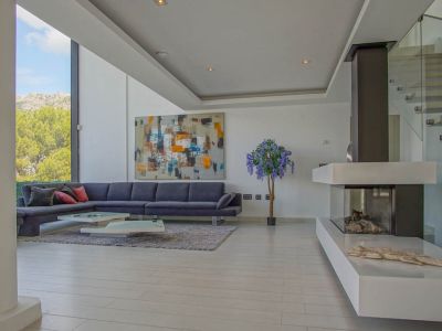 Geräumiges Wohnzimmer mit grauem Ecksofa, modernen Couchtischen und Gaskamin. Große Fenster bieten Berg- und Baumblick, ergänzt durch eine Glastreppe.